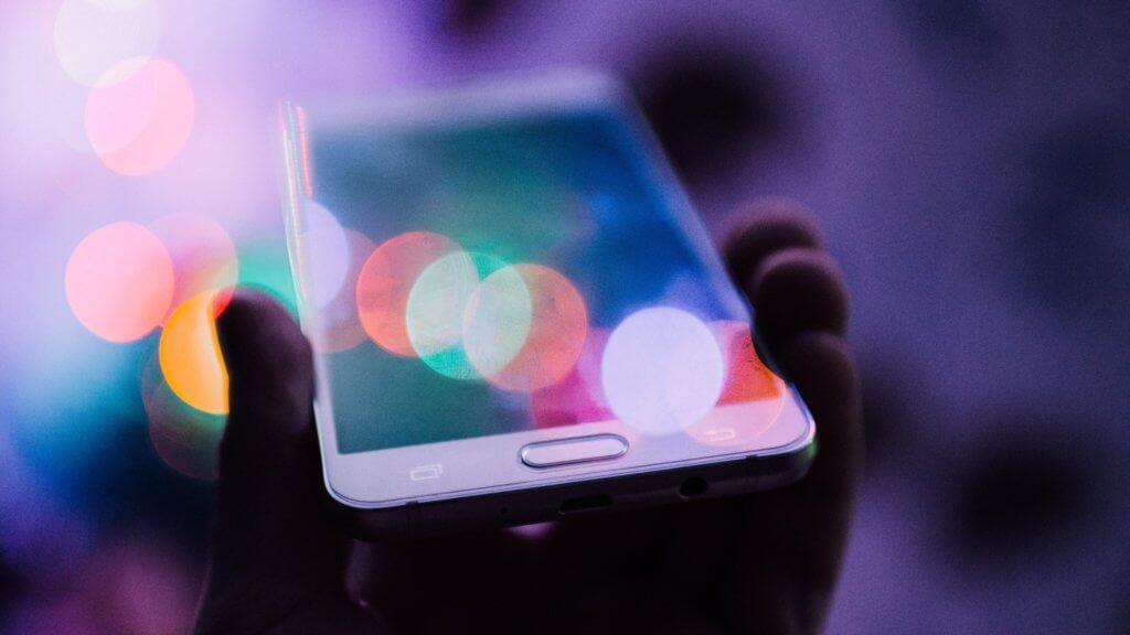 Photograph of a hand holding a mobile phone with blurred lights reflecting off the screen.
