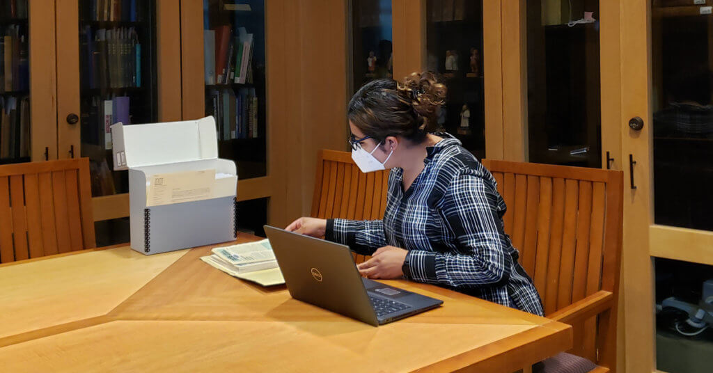 A person wearing a face mask sitting a table using a laptop computer.