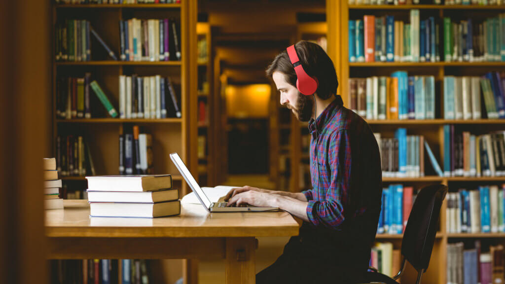 A man wearing red headphones sits at a table in a library working on a laptop surrounded by books