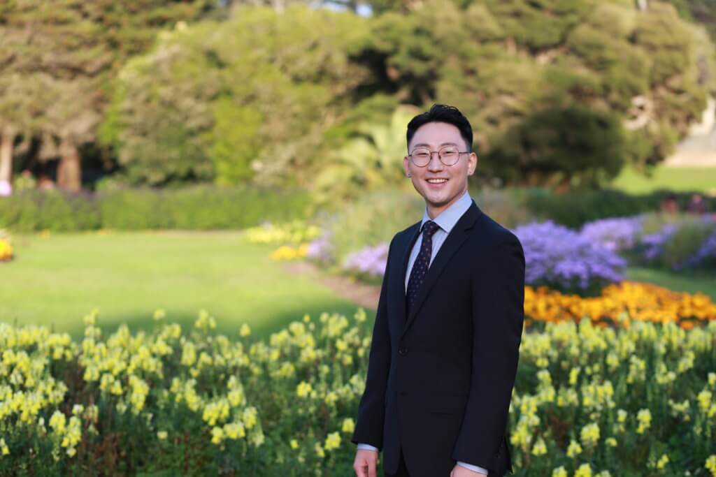 James Lim, a UCSF MD, MAS student dressed in a suit and tie. In the background are trees, grass and groups of yellow, orange and purple flowers