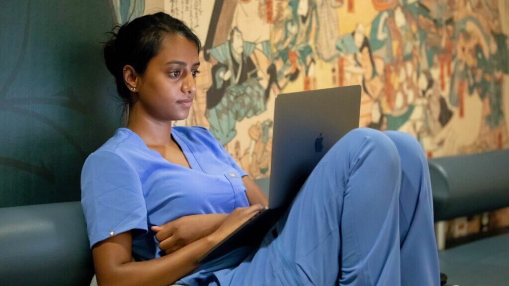 Student in blue scrubs sitting with laptiop propped on their knees. The wall behind the student contains images from the UCSF Japanese Woodblock collection.
