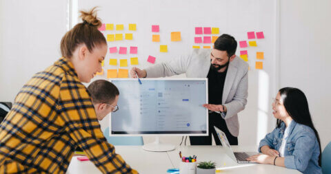 Group of four people working together at a desk, referencing information on a screen. In the backfround is a wall full of post-it notes