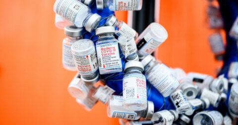 Empty bottles of COVID-19 vaccine hang on a blue structure with an orange wall in the background