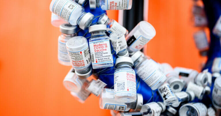 Empty bottles of COVID-19 vaccine hang on a blue structure with an orange wall in the background