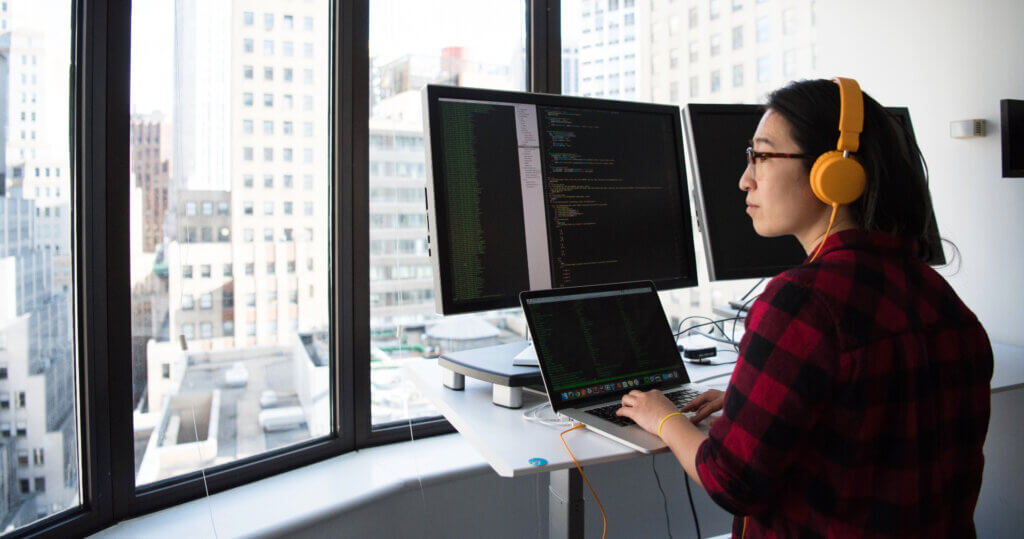 Person working at a standing desk facing two computer monitors, looking at code. They are wearing orange headphones and a red flannel.