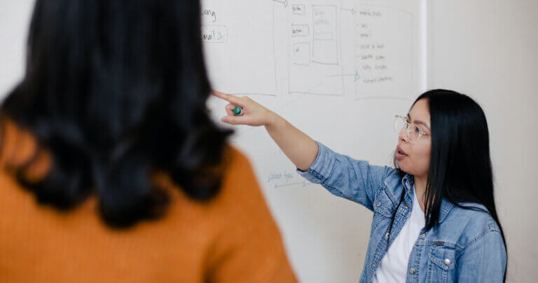 Woman with long black hair wearing a denim jacket pointing to information on a white board and explaning to her colleague in an orange sweater.