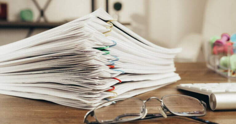 Documents held together by colorful paper clips stacked together on a desk, in the foreground is a pair of reading glasses.
