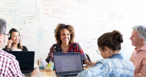 Five people are sitting at a table facing each other, two people have laptops. On the wall behind the group is a whiteboard with writing on it.