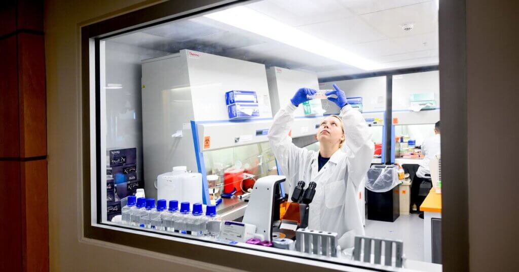 Researcher in a lab looking up while holding a sample with gloved hands.