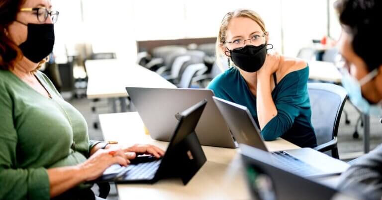 Three people sitting together, wearing masks and working on laptops