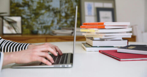 Hands on a laptop in the background are books stacked on the same table