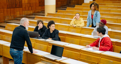 Six students and a professor in a large lecture hall with rows of seating.