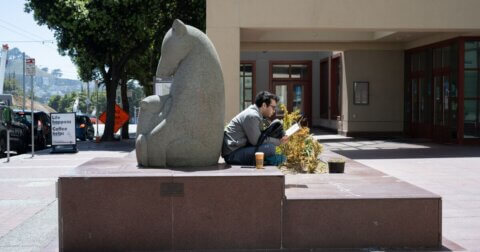 Person reading and sitting with their back touching a granite sculpture of a mother bear nursing her young cubs in front of the Kalmanovitz Library.