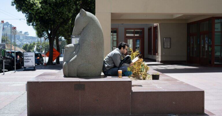 Person reading and sitting with their back touching a granite sculpture of a mother bear nursing her young cubs in front of the Kalmanovitz Library.