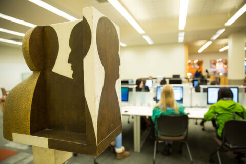 Art piece on the main floor of the Kalmanovitz Library with profiles of a face in different directions, in the background are people seated at desktop computers.