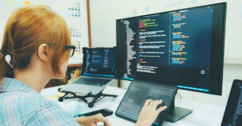 A person seated at a computer desk in front of a monitor that shows coding, a laptop on a stand is to their left and a tablet is in front of them.