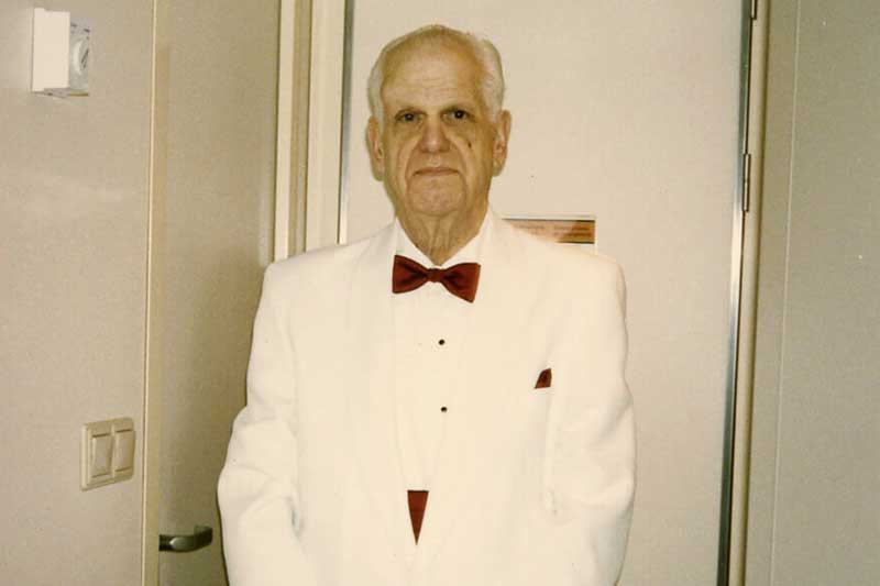 Eli Hassid MD, in a white tuxedo with red bow tie, standing indoors.