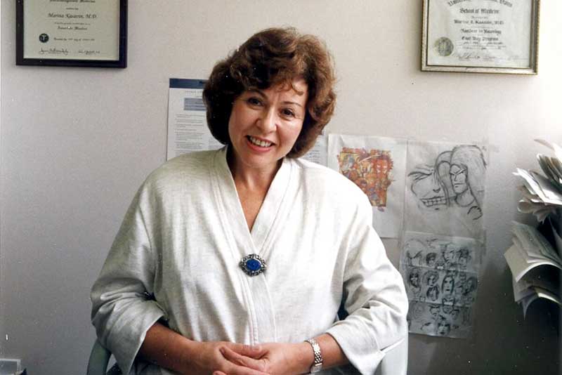 Marina Kasavin, MD, in a cream blouse with a blue brooch sits smiling in an office, with framed certificates and sketches on the wall behind her.