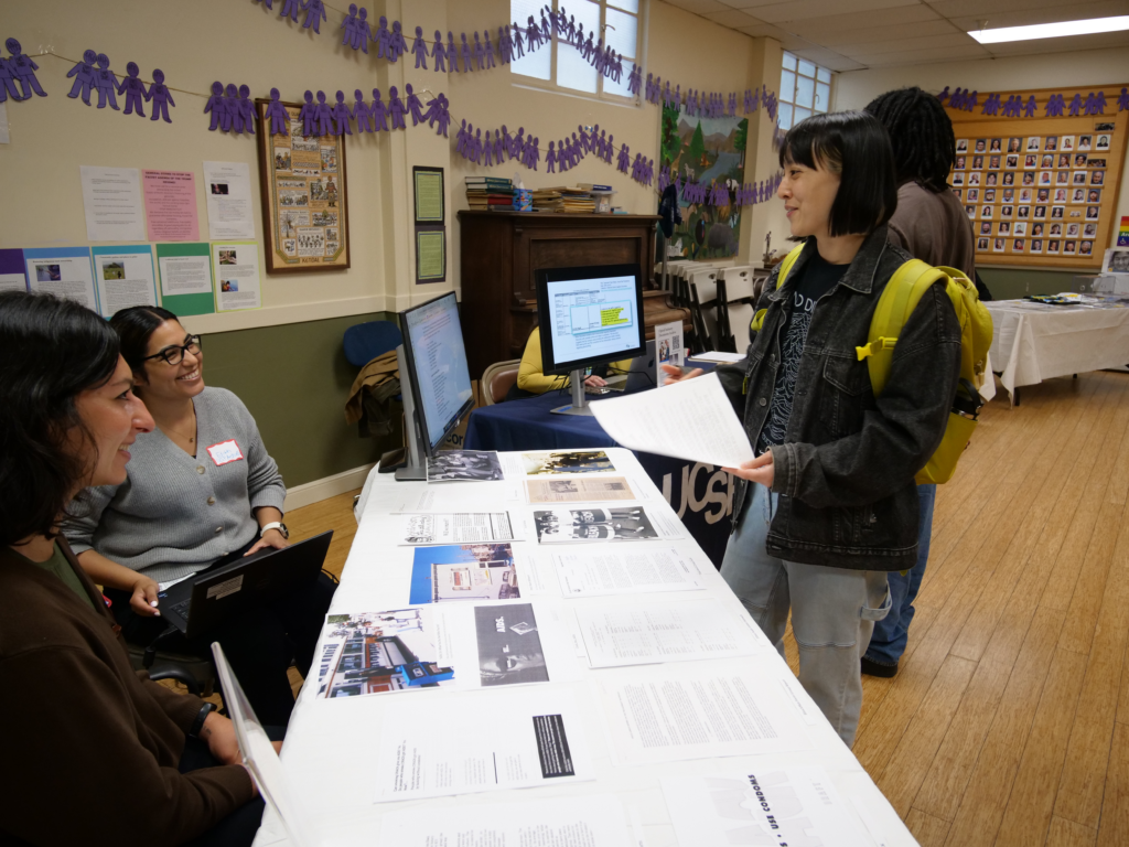 Two archivists smiling and seated a table inside speaking to a person wearing a yellow backpack and holding a paper.