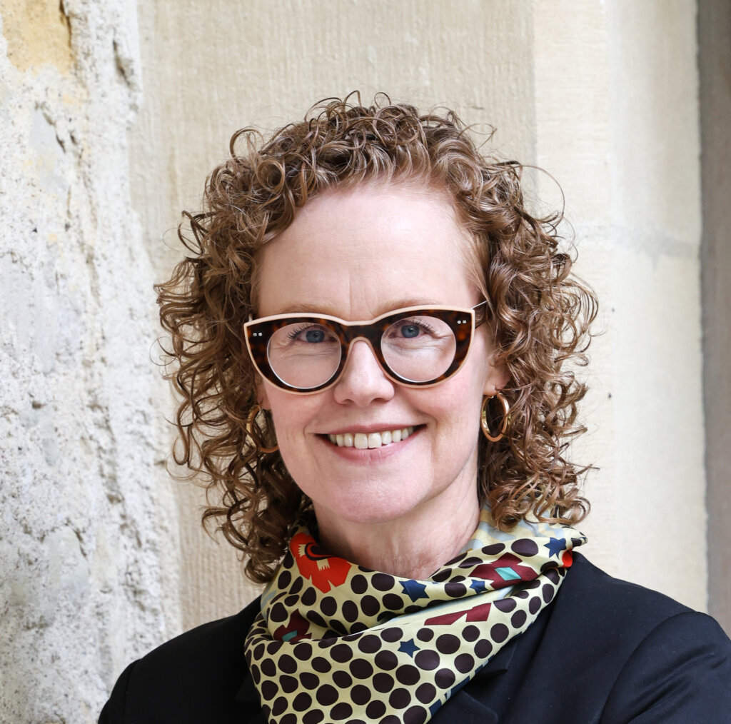 Kristen Ostherr, smiling, wearing tortoise shell framed glasses and a colorful scarf with a vibrant pattern.