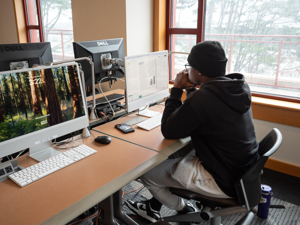 Student seated at a work station near a window and looking at a computer monitor in the library computer classroom CL 230.