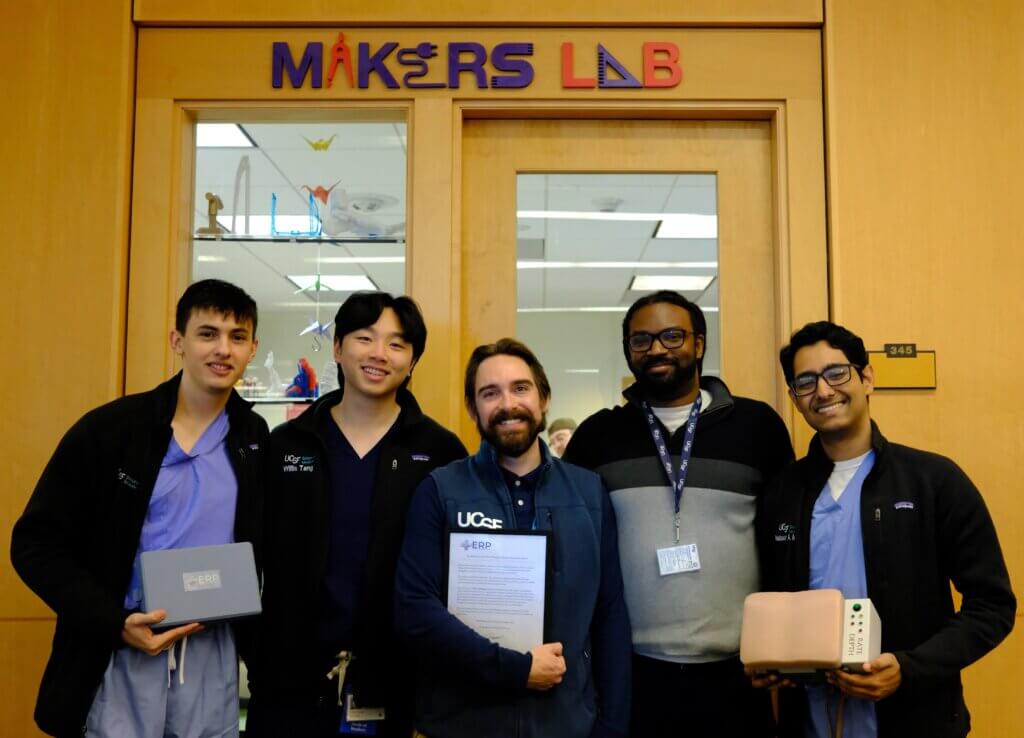 Three medical school students and two Makers Lab engineers standing together, smiling and holding components of the CPR model in front of the Makers Lab at UCSF.