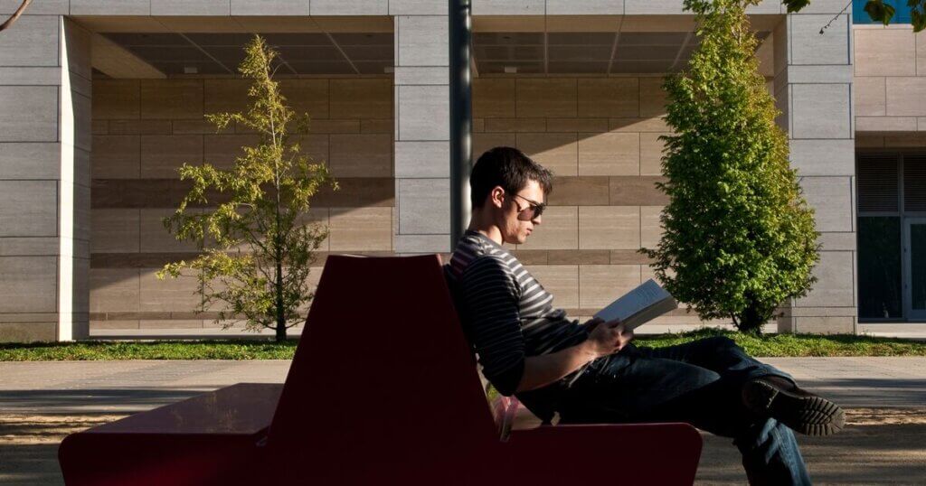Person wearing sunglasses seated outside on a bench and reading a book.