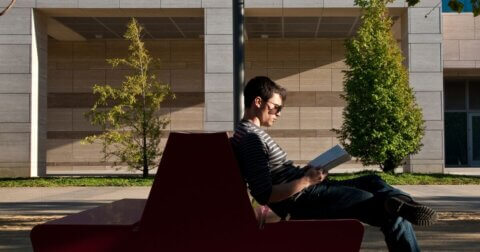 Person wearing sunglasses seated outside on a bench and reading a book.