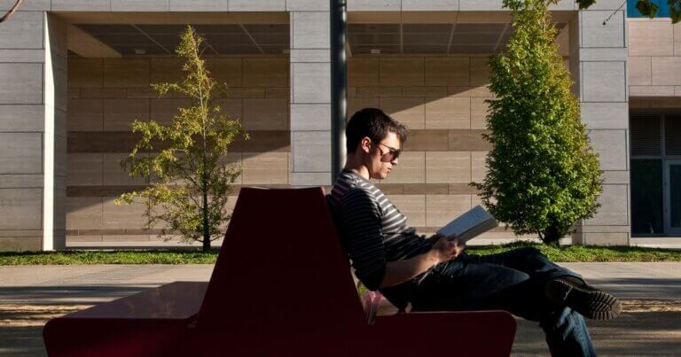 Person wearing sunglasses seated outside on a bench and reading a book.