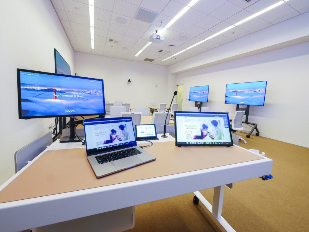 Standing desk with a microphone, monitor screen, laptop screen, and two tablet screens with a white walled classroom space in the background.