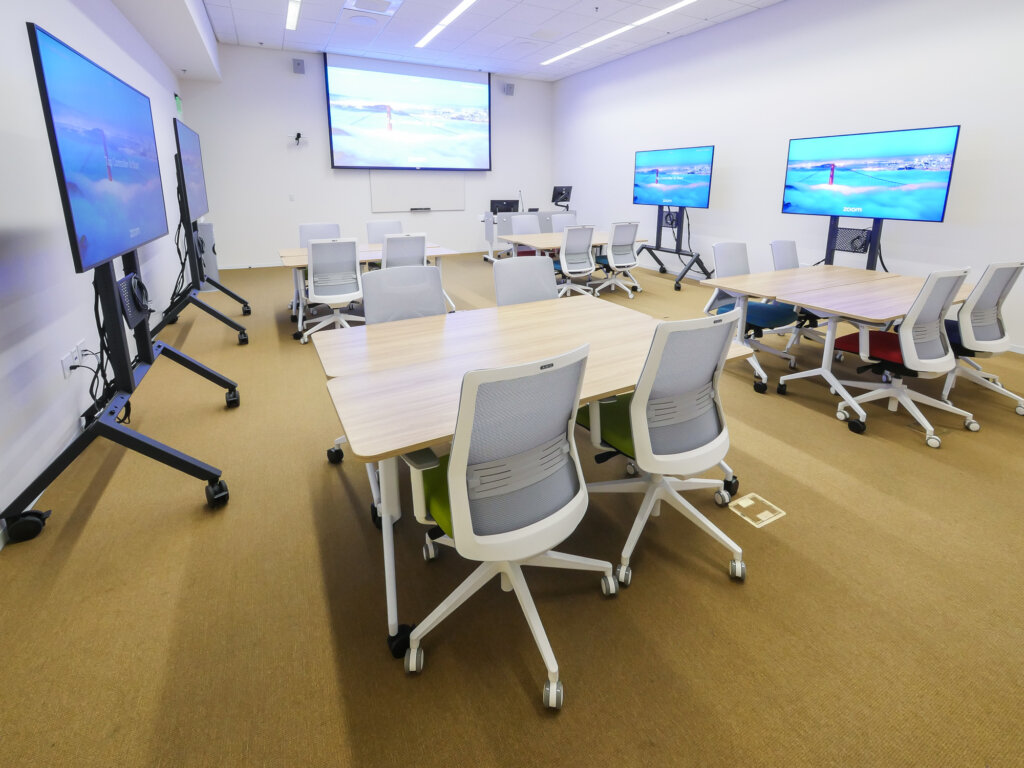 A white walled classroom with a projector screen on the main wall, four tables with four desk chairs around each, four large black screens on wheels against the walls.
