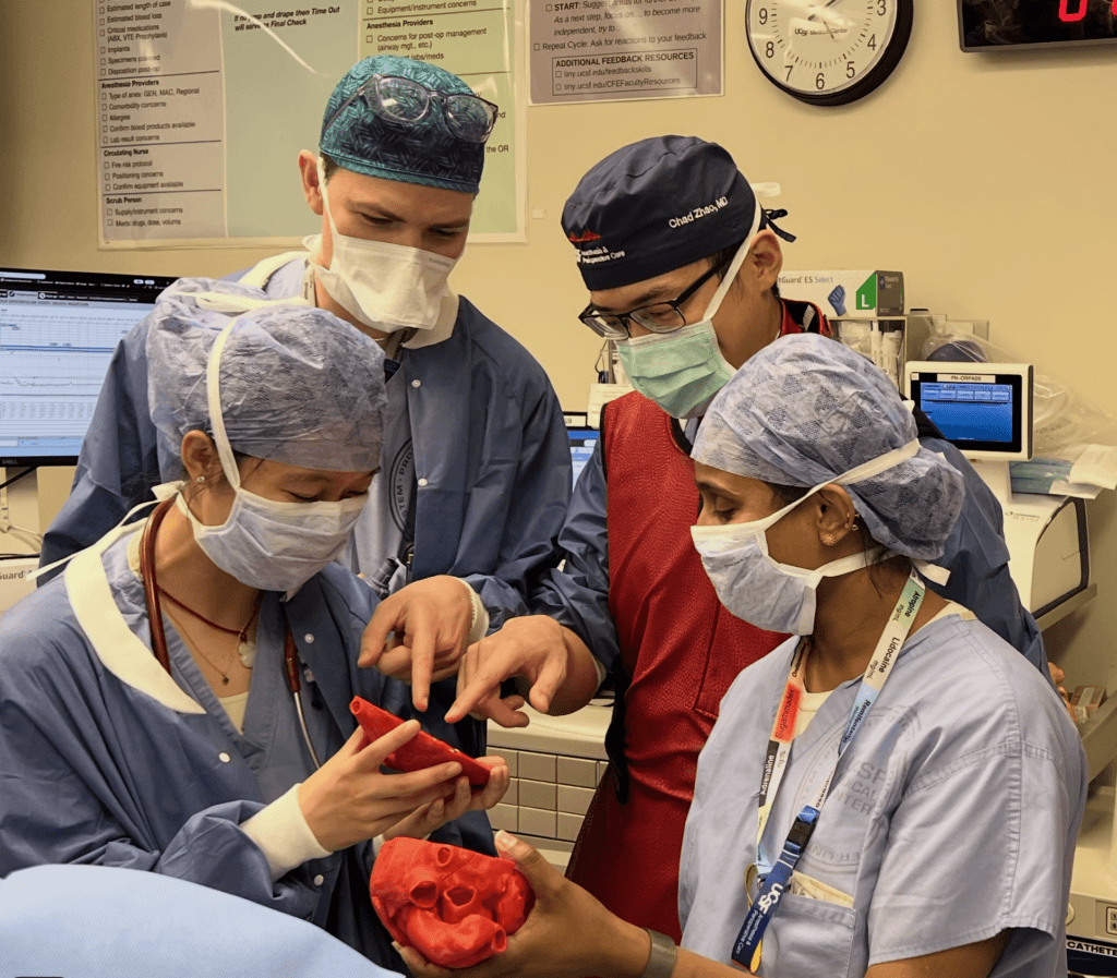 Litty John and her 3 fellows in masks and scrubs looking down to examine a 3D printed heart model in the OR