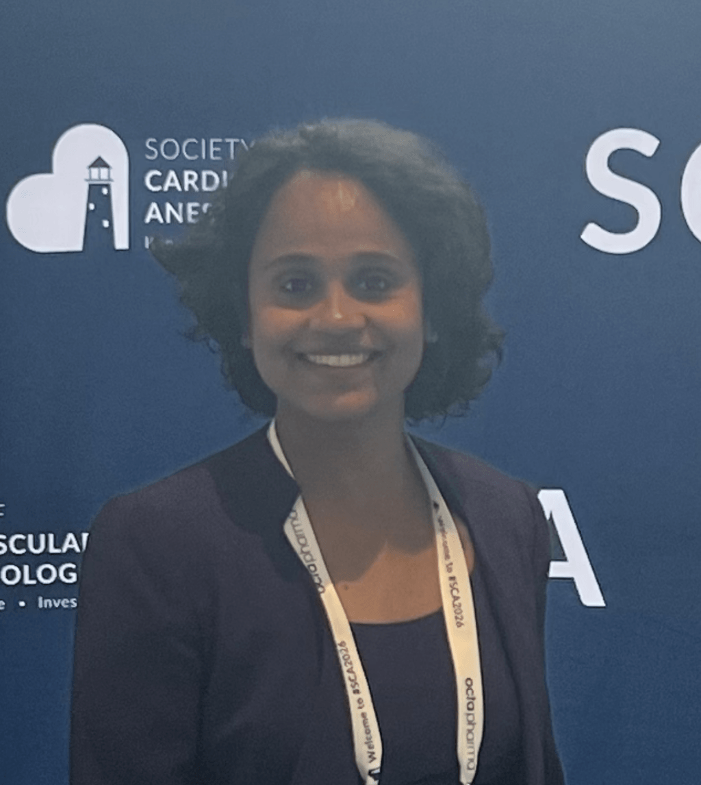 Litty John, smiling and standing in front of Society of Cardiovascular Anesthesiologists (SCA) banner