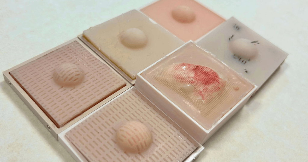 Close up photo of 6 square shaped skin condition trays holding silicone cyst and lipoma models of varying condition.