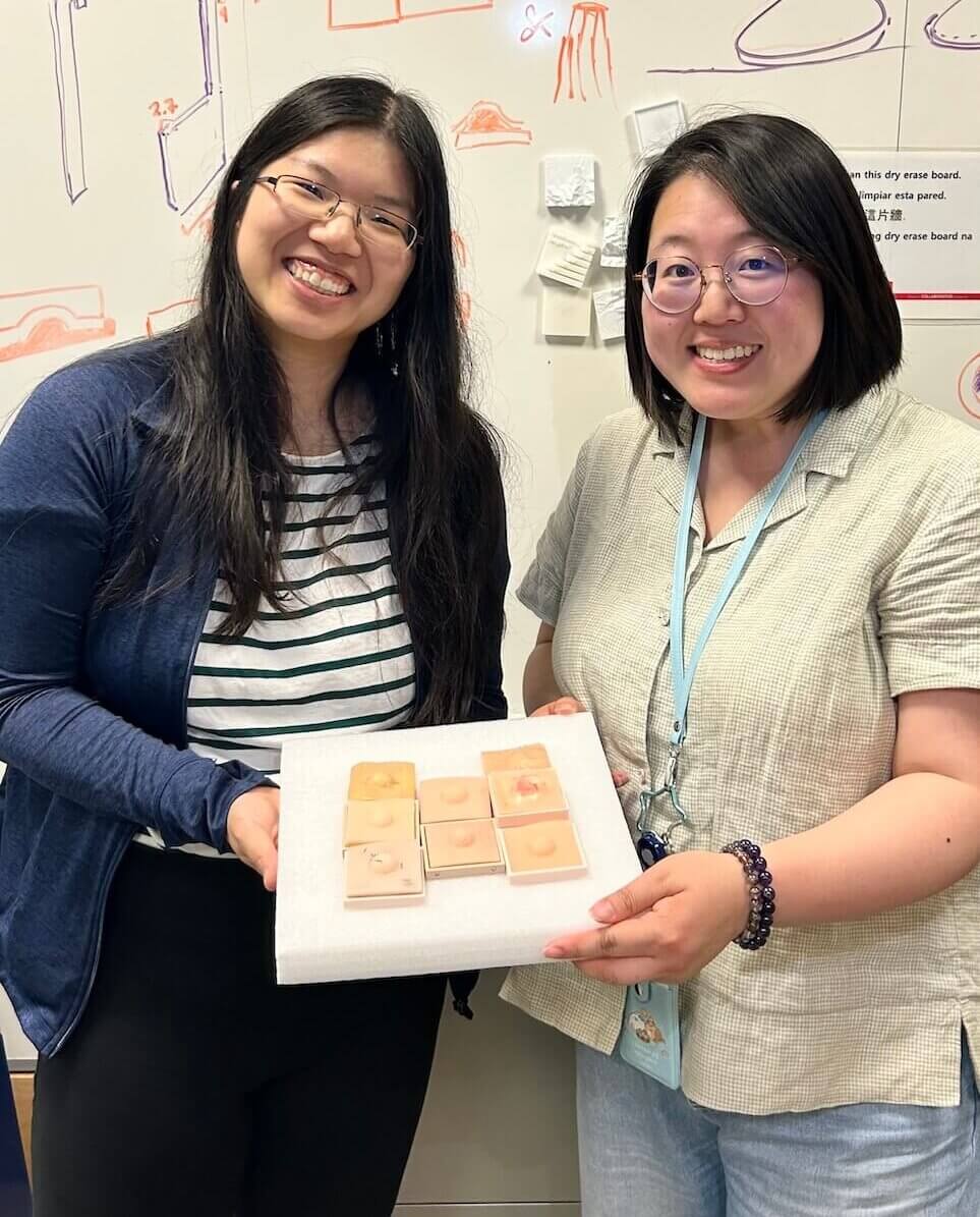 Alice Tang, MD, PhD, displays a tray of sample skin conditions with Jenny Tai, Makers Lab engineer, in front of a whiteboard.
