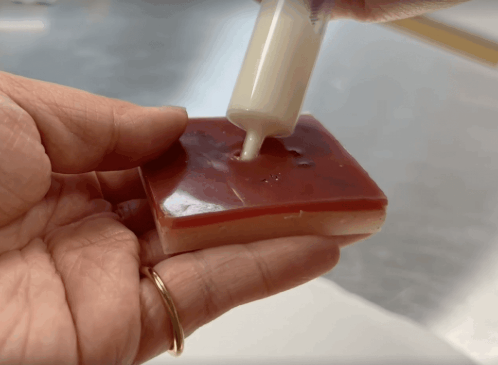 Close up of a hand holding a square shaped silicone block and using a syringe to inject lotion into the opening.