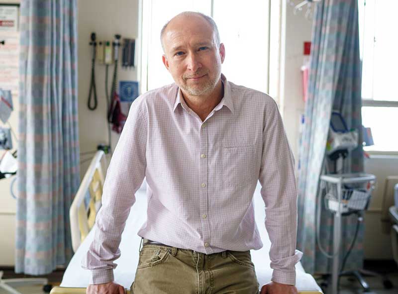Steven Deeks, MD, seated upright at the end of a hospital bed in a hospital room.